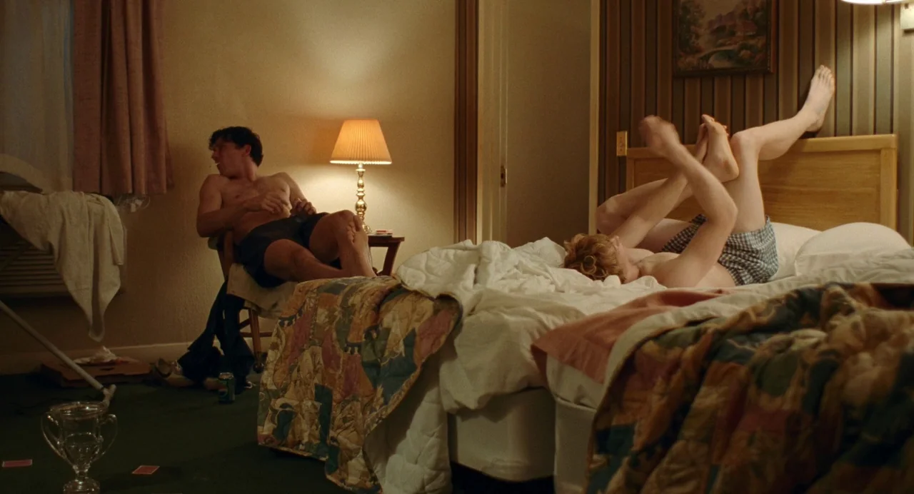 Challengers (2024) movie still: wide — A dated motel room features two beds with floral patterned quilts and dark wall paneling. One man sits on a…