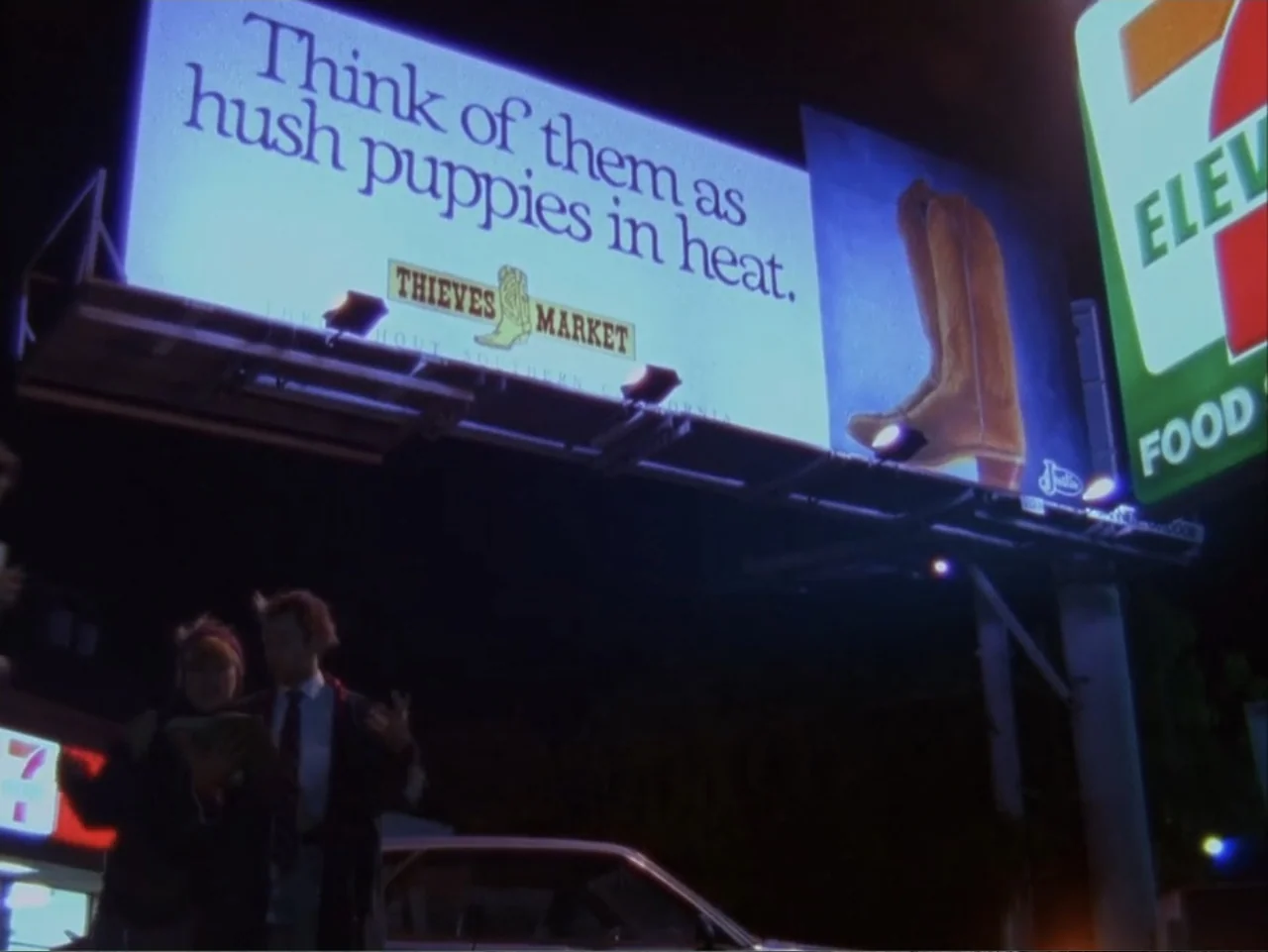 The Living End (1992) movie still: medium — A night scene outside a gas station featuring a large billboard advertisement above. Two people stand…