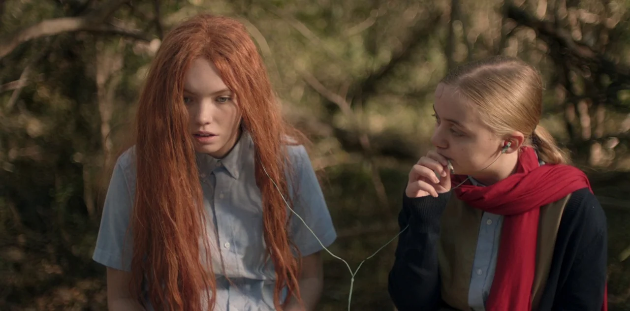 Darlin’ (2019) movie still: medium — Two teenage girls sit in a wooded area with trees blurred in the background. They share a single pair of wired…