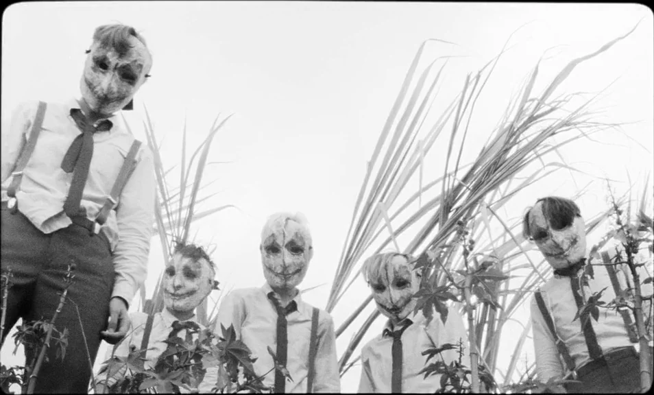 The Wild Boys (2017) movie still: fullbody — Five figures in identical dress and masks stand in a tall grass field. The camera looks up from below…