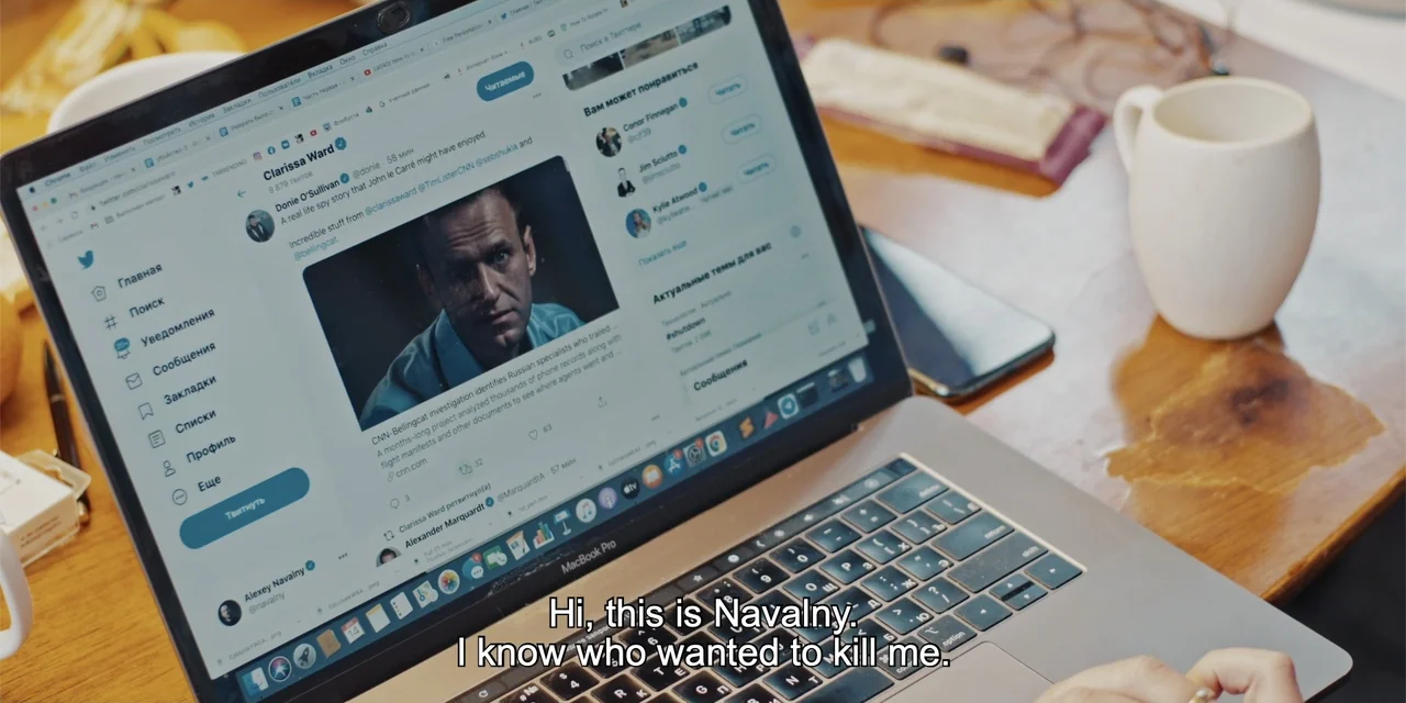 Navalny (2022) movie still: closeup — A wooden desktop surface holds a laptop displaying a social media profile page. A person's hands are visible…
