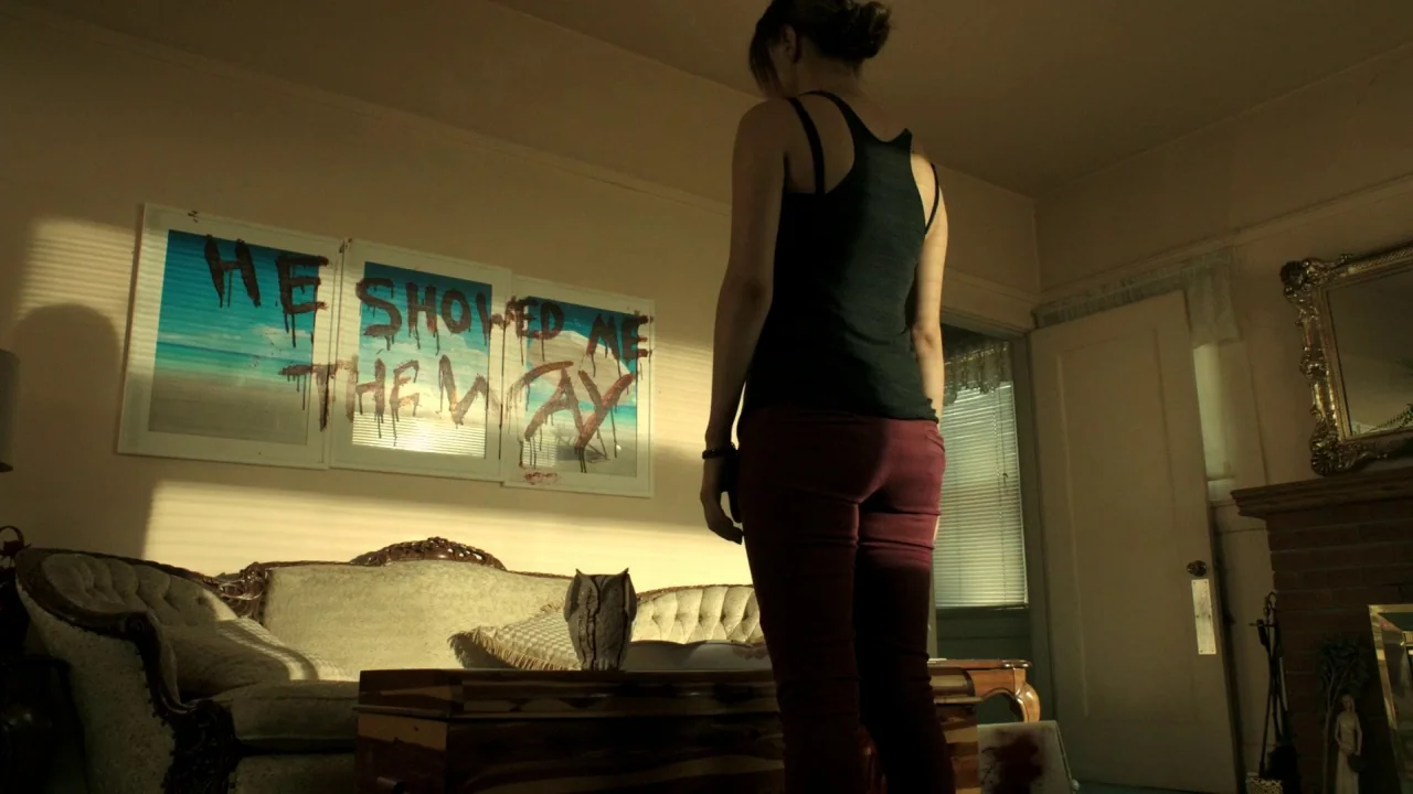 The Pact II movie still: medium — A furnished interior room features a couch against a wall covered in dark text. A woman stands in the foreground…