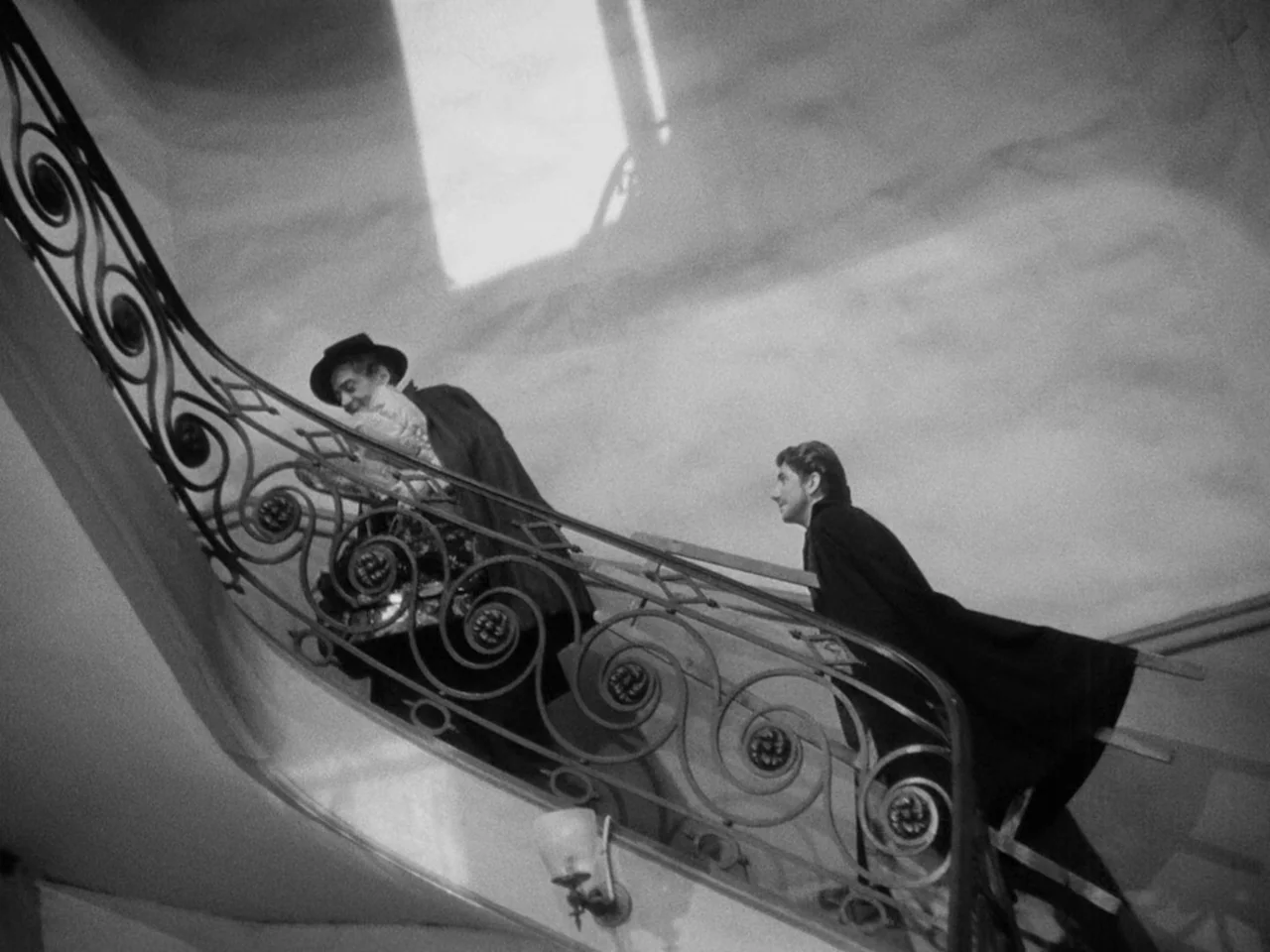 Le Plaisir (1952) movie still: medium — An ornate interior staircase features curved metal railings against a plain high-ceilinged wall. Two men in…