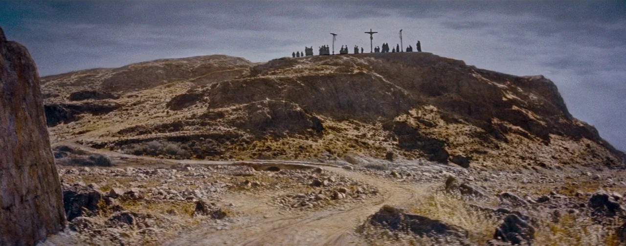 The Robe (1953) movie still: establishing — A rocky, arid hill rises under a heavy, overcast grey sky. Silhouetted figures stand on the peak,…