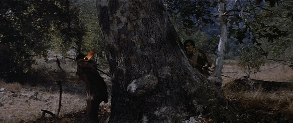 Flaming Star (1960) movie still: wide — A dense forest with dry grass and a massive, textured tree trunk center frame. Two men positioned on either…