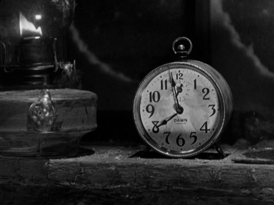 The Gold Rush (1925) movie still: closeup — An analog alarm clock sits on a dusty, wooden surface next to an oil lamp. The clock face shows the…