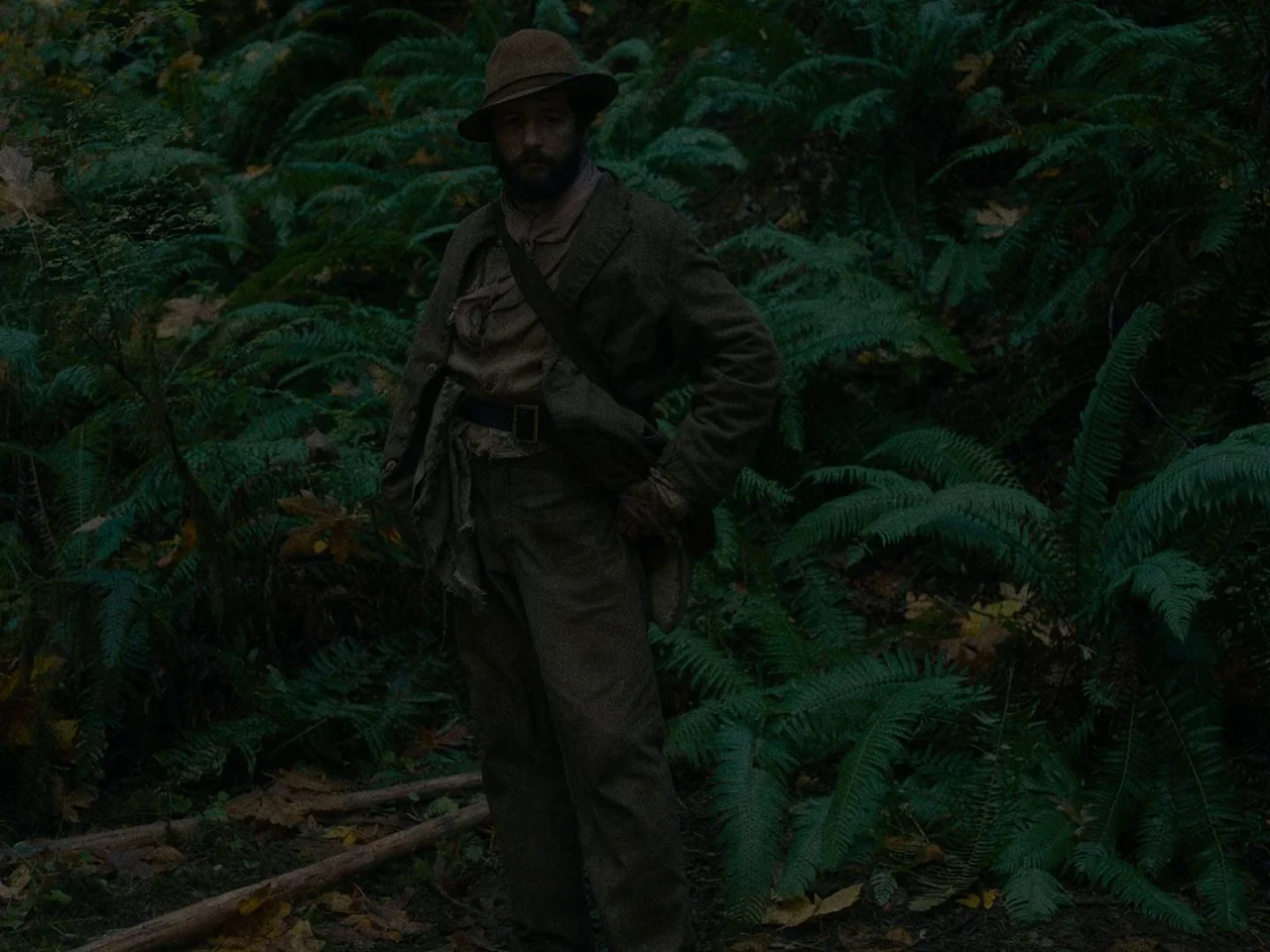 First Cow (2019) movie still: fullbody — A man stands in a dense, dark temperate rainforest overgrown with lush green ferns. The subject remains…
