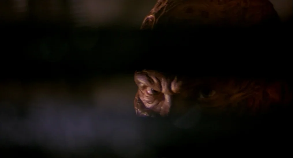A Nightmare On Elm Street (1984) movie still: closeup — A dark bedroom interior with a character hidden mostly by dense shadows. The subject stares…