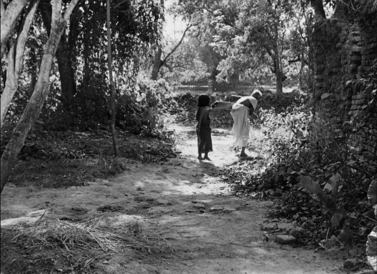 Pather Panchali (1955) movie still: wide — A rural earthen path winds past a crumbling brick structure and dense foliage. An elderly woman and a…