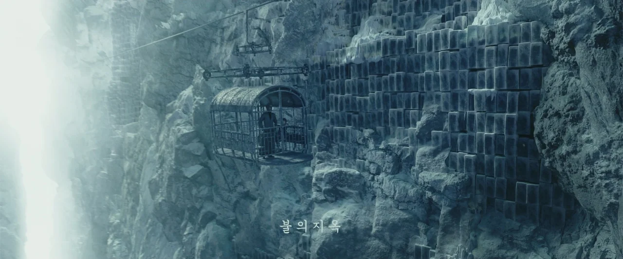 Along with the Gods: The Two Worlds (2017) movie still: wide — A metal cage hangs from a cable against a towering, sheer stone cliff face. A man…