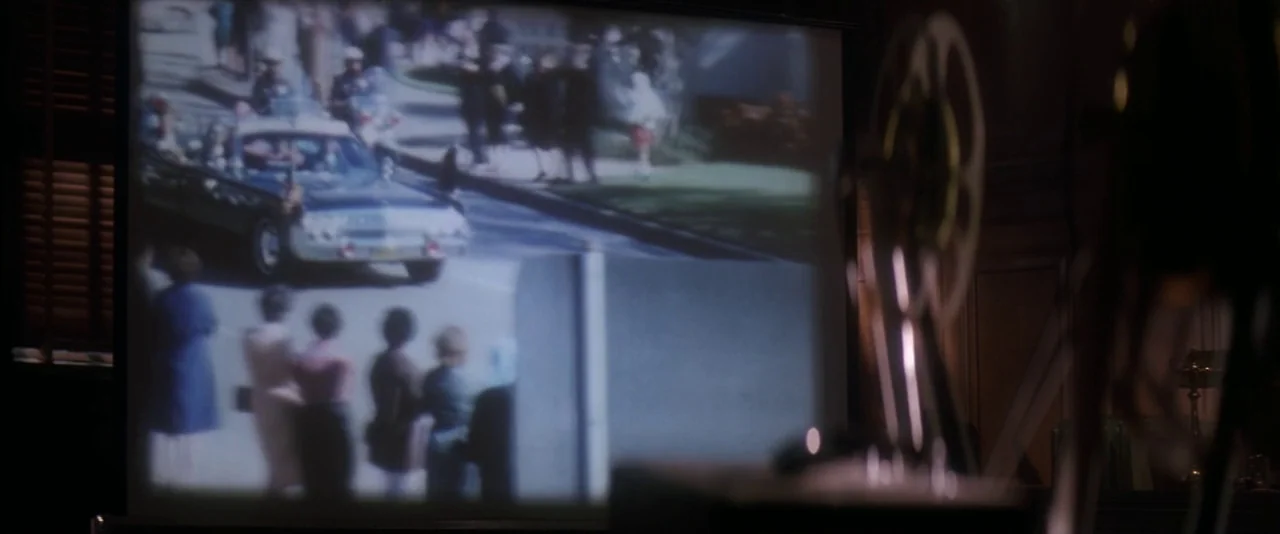 JFK (1991) movie still: medium — A dark projection room features a screen displaying a 1960s motorcade with spectators. The mechanical projector…