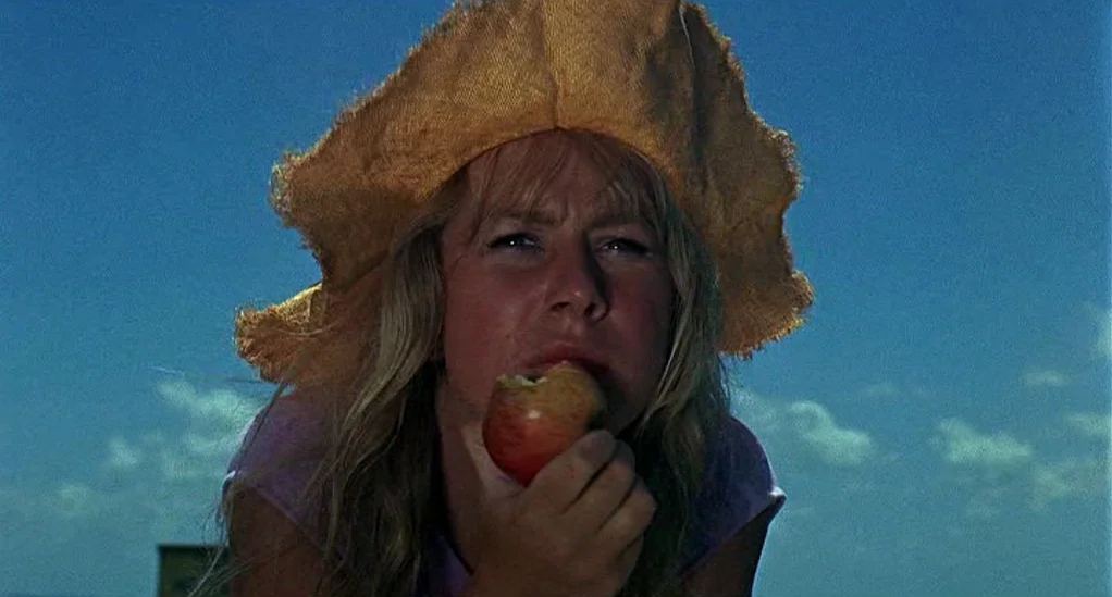 Age of Consent (1969) movie still: closeup — A woman wears a large straw hat against a clear bright blue sky. She holds a red apple to her mouth…