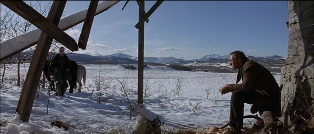 Unforgiven (1992) movie still: wide — A dilapidated, partially collapsed wooden structure sits on a vast, snow-covered mountain landscape. A man…