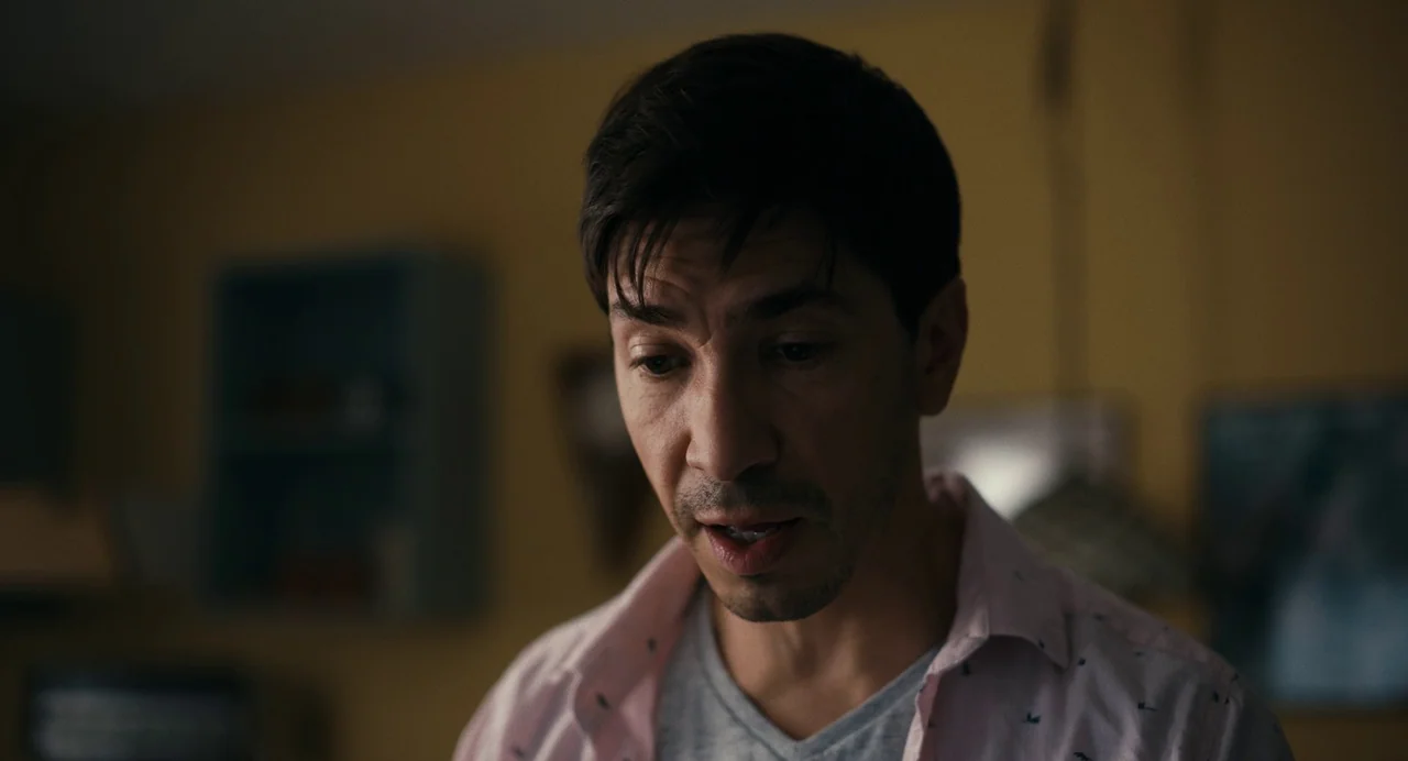 Barbarian (2022) movie still: closeup — A man stands indoors against a muted yellow wall with out-of-focus household objects behind. The subject is…