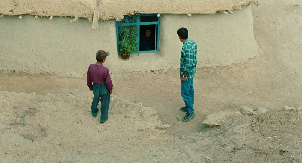 The Wind Will Carry Us (1999) movie still: wide — A rural earthen structure with a blue window frame sits on a dusty slope. Two people stand in the…