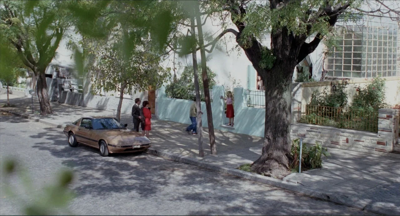 Waiting for the Hearse (1985) movie still: wide — A sunny residential street features a tan sports car parked by a wall. A small group of people…