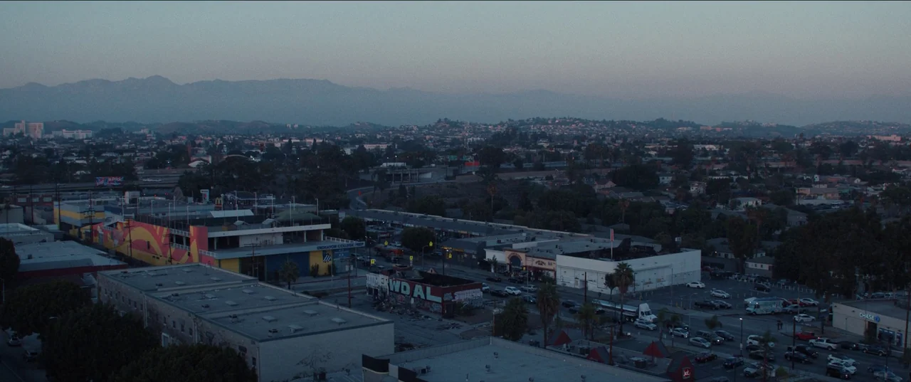The Greatest Hits (2024) movie still: establishing — An aerial panoramic view of a densely populated city sprawl under a twilight sky. Low-rise…
