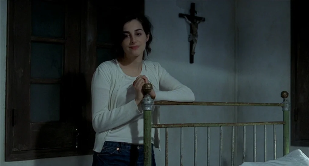 Anatomy of Hell (2004) movie still: medium — A spartan room features a green metal headboard against a white wall. A woman stands by the bed while a…
