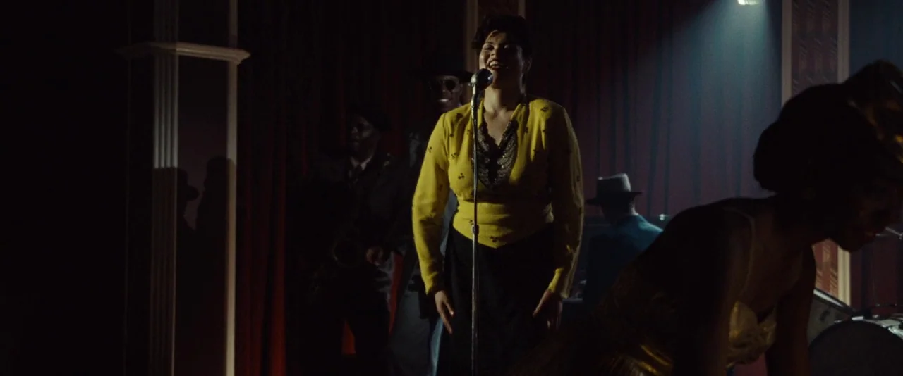 Kill Your Darlings (2013) movie still: medium — A dimly lit nightclub stage features deep red curtains and wood paneled pillars. A woman stands…