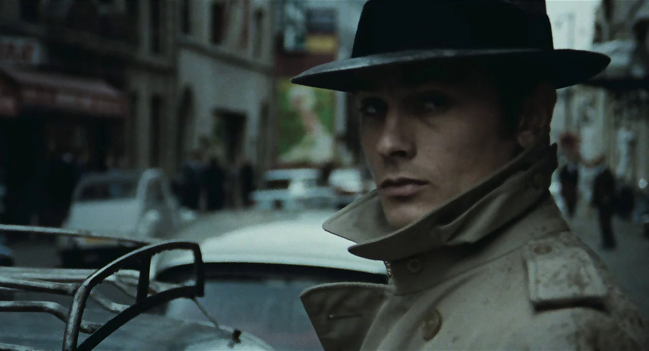 Le Samouraï (1967) movie still: closeup — A city street with blurred buildings and parked cars fills the background. A man stands in the foreground…