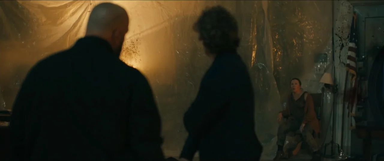 Guns Up (2025) movie still: medium — A dimly lit room with plastic sheeting covering the walls and an American flag. Two individuals in dark…