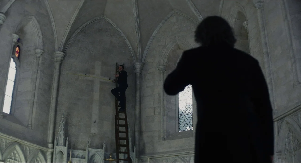 Wake Up Dead Man (2025) movie still: wide — A stone cathedral interior with ribbed vaults features a high, light-colored wall. A man in black stands…