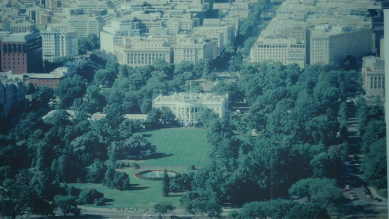 Demon in the White House (2021) movie still: establishing — A high-altitude aerial view of a large white building centered within a grassy field.…