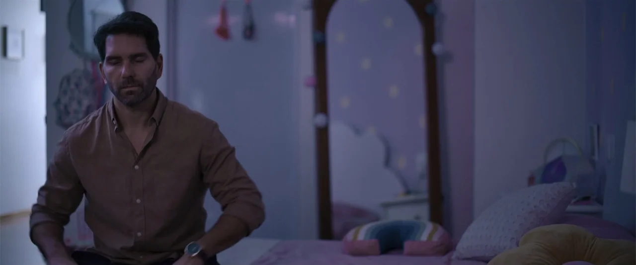Evil Eye (2020) movie still: medium — A dimly lit child's room features a bed with soft pillows and a mirror. A man in a tan shirt sits on the bed…