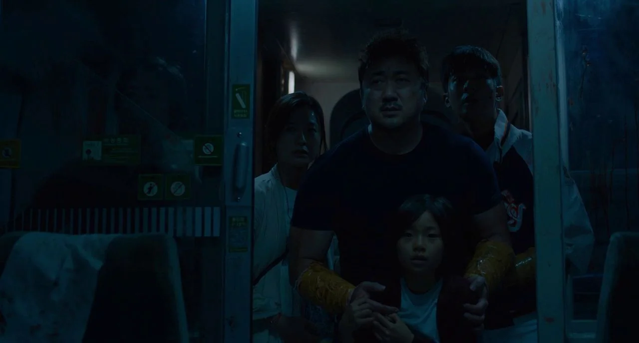 Train To Busan movie still: medium — A dimly lit train carriage features a metal door framing a group of people. The man stands centrally, shielding…
