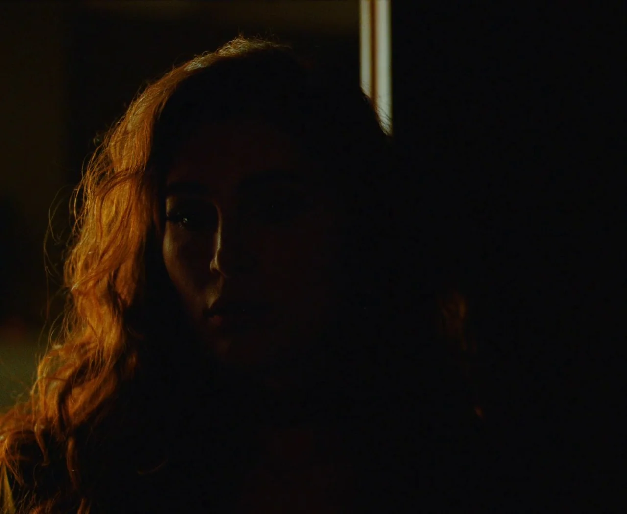 Monica (2022) movie still: closeup — A dimly lit indoor space with shadows concealing most of the environment. A woman with long wavy hair has one…