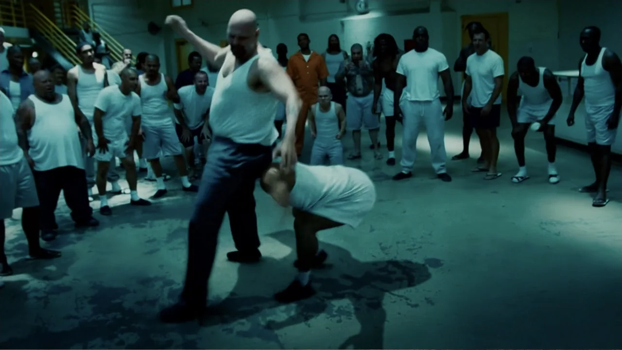 Hancock (2008) movie still: fullbody — A large concrete prison dayroom contains a crowd of men in various white uniforms. A bald man in a tank top…