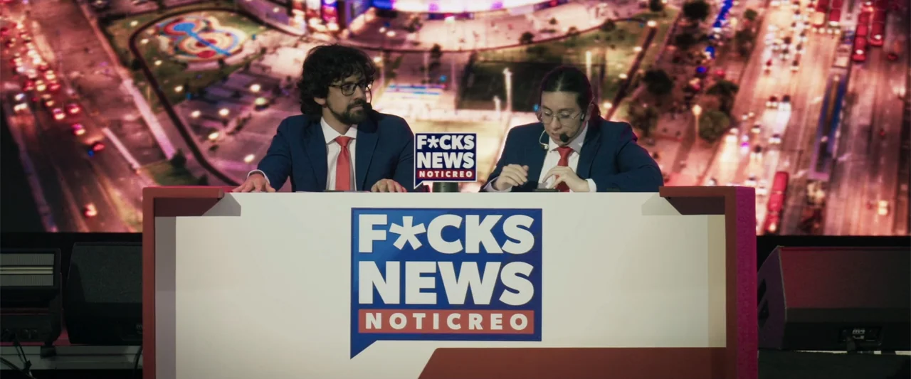 Making of: F*CKS NEWS (2025) movie still: medium — A news studio set features a large white desk with prominent blue and red signage. Two male…