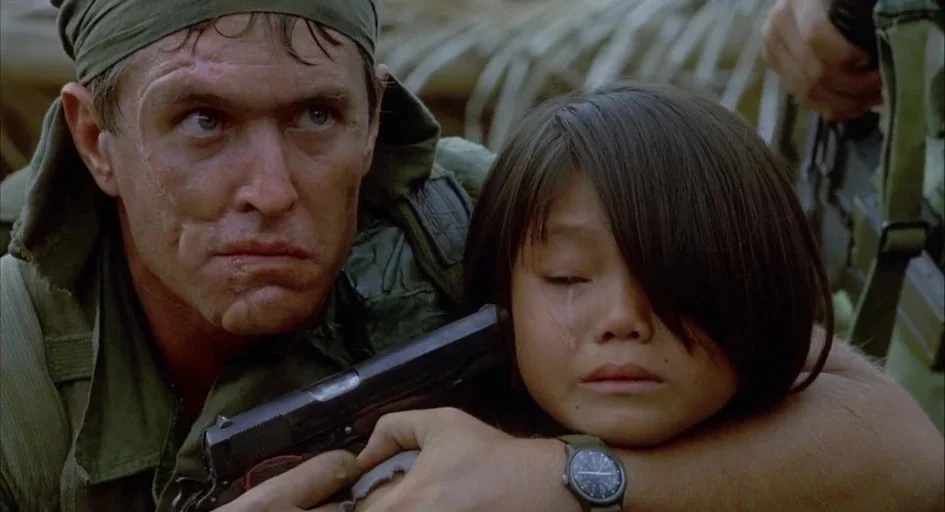 Platoon (1986) movie still: closeup — A soldier wearing a headband holds a pistol near a young child's temple. The background is blurred with muted…