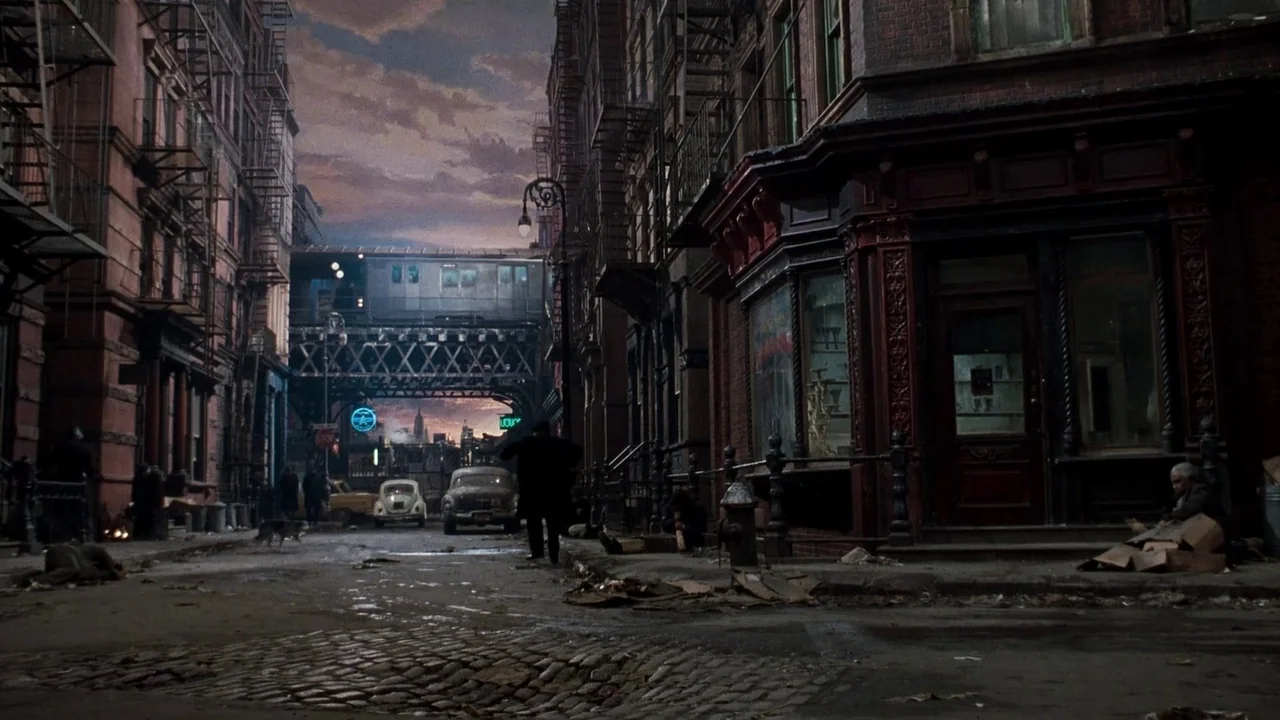 Little Shop of Horrors (1986) movie still: wide — A dense urban intersection features dark brick tenements with rusted iron fire escapes. A lone man…