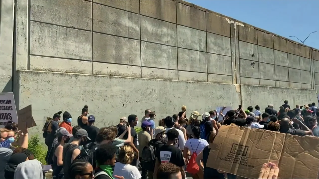 Covid Diaries NYC (2021) movie still: wide — A dense crowd of people gathers along a tall weathered concrete retaining wall. The individuals stand…