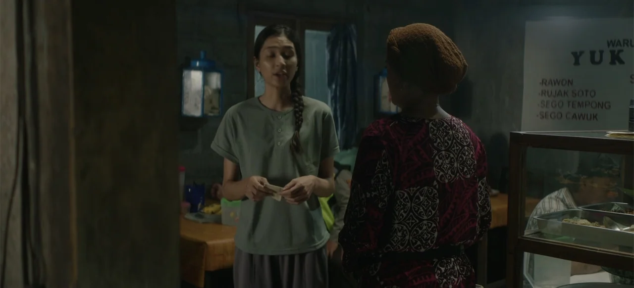 A Thousand Days (2023) movie still: medium — A dimly lit indoor food stall features a menu sign and food display counter. Two women face each other…