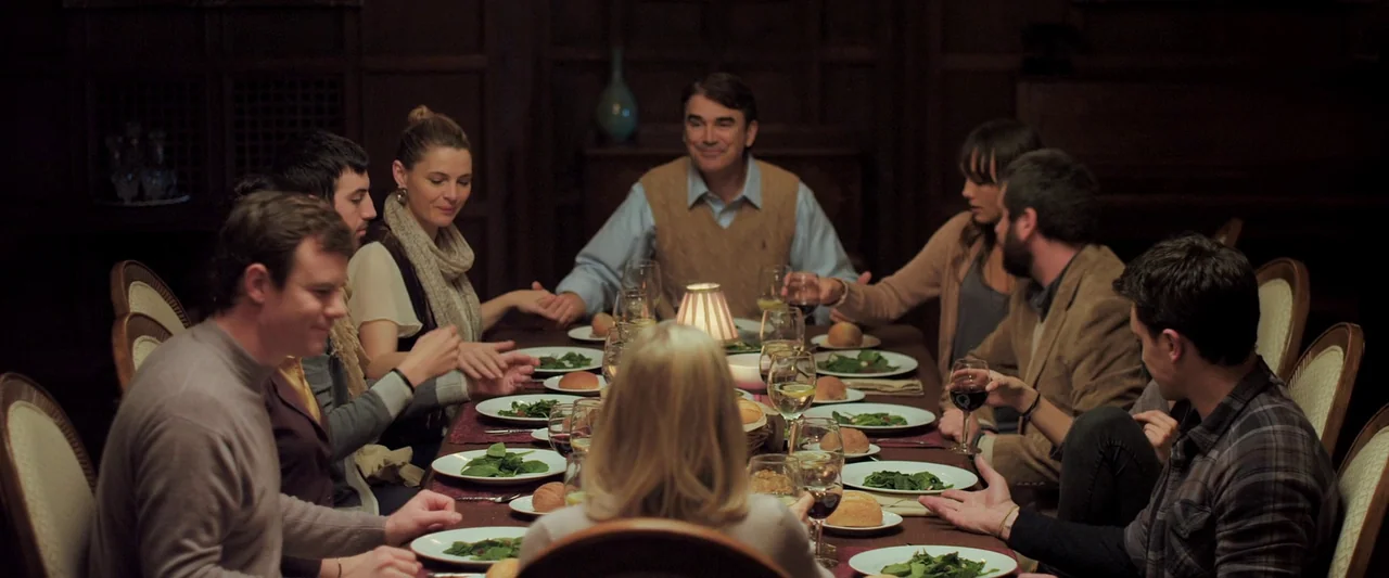 You’re Next (2013) movie still: wide — A group of people sits around a long wooden table in a dimly lit room. They hold hands and look toward the…