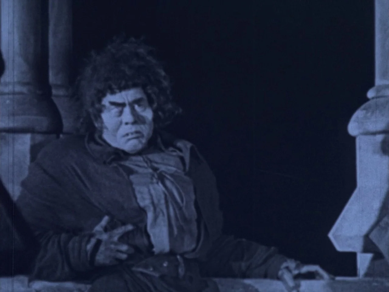 The Hunchback of Notre Dame (1923) movie still: medium — A stone balcony at night with architectural columns flanking the frame. A man with a…