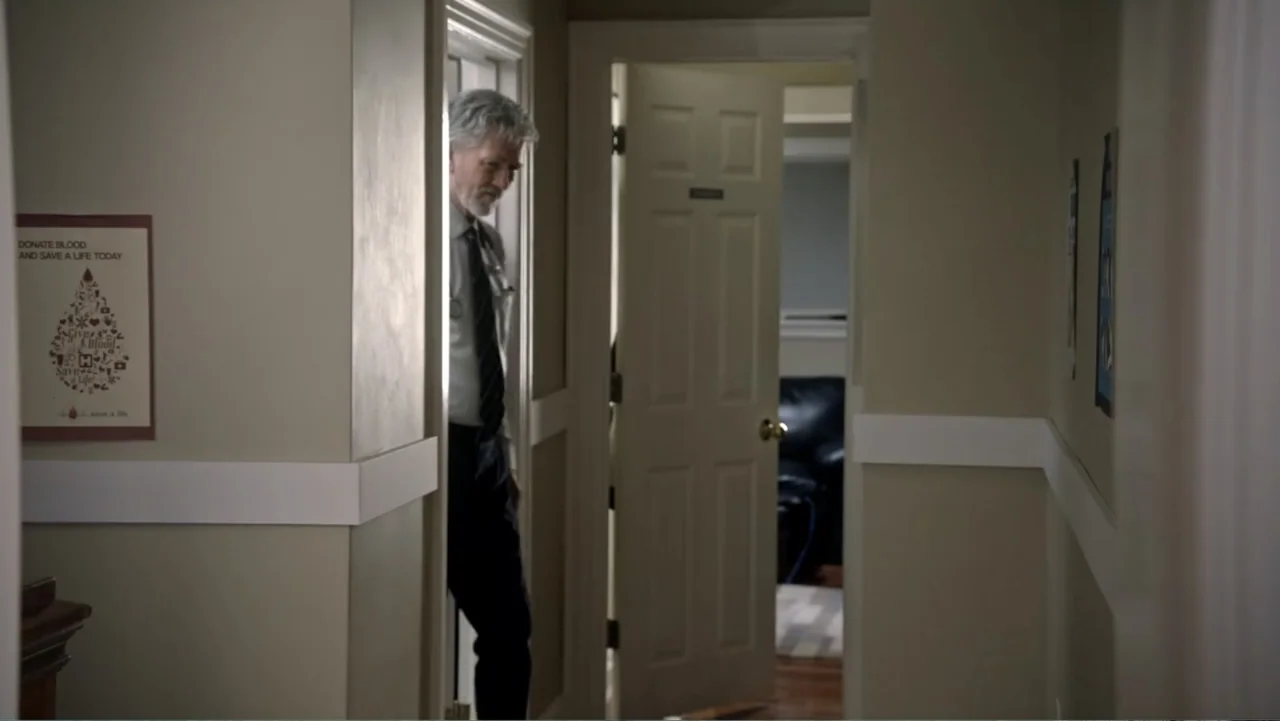 The Christmas Cure (2017) movie still: medium — A quiet indoor hallway with beige walls and a blood donation poster on one side. A middle-aged man…