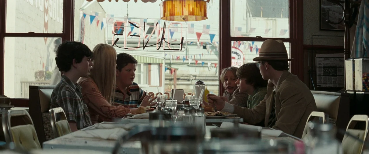 Super 8 (2011) movie still: wide — A group of six teenagers sit in a vinyl booth inside a sunlit restaurant. They are sharing a meal while…