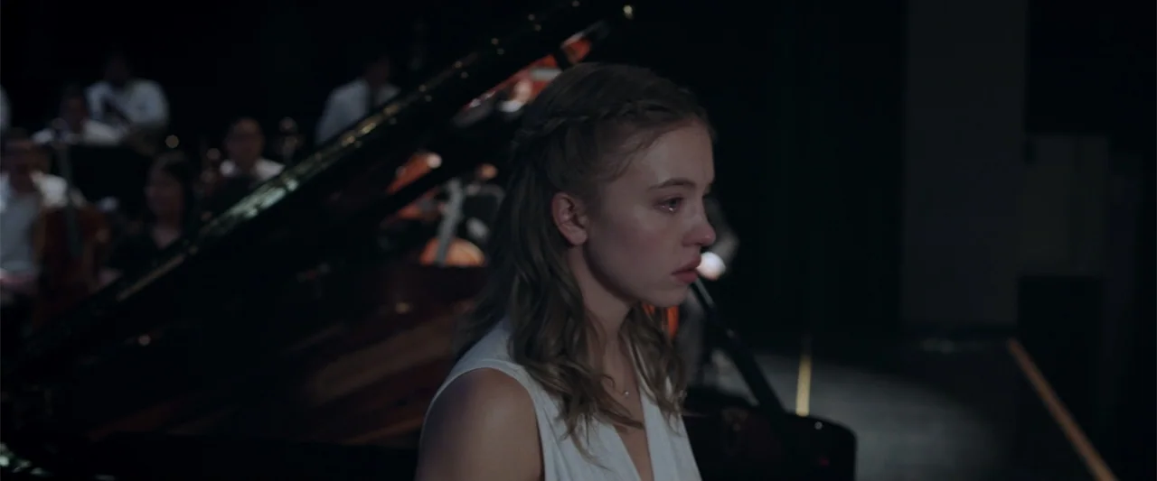 Nocturne (2020) movie still: medium — A dark concert hall interior with a grand piano in the background. A young woman in a white top sits at the…