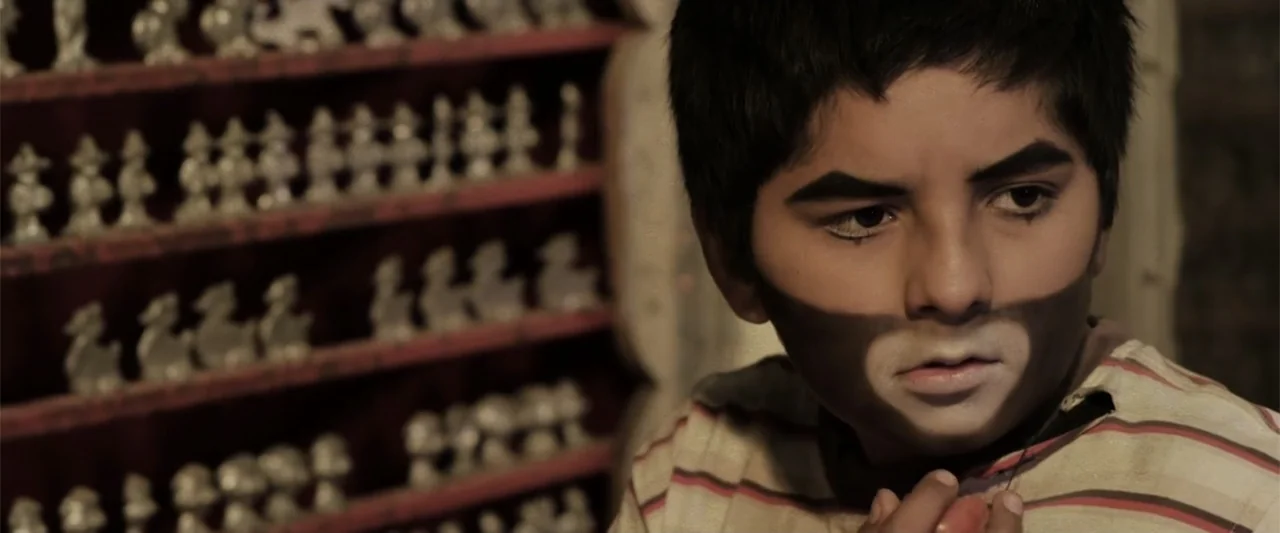 At Dawn (2013) movie still: closeup — A wooden shelf holding rows of small decorative figures stands behind a young boy. The boy wears theatrical…