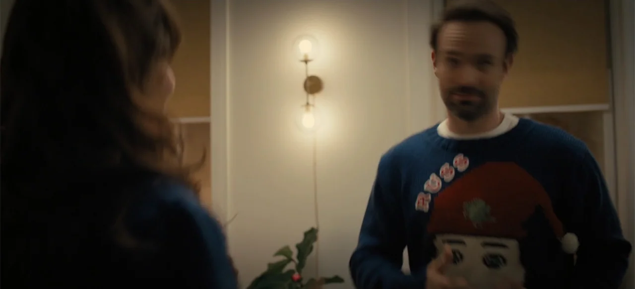 Merv (2025) movie still: medium — A man in a Christmas sweater stands in a dimly lit home interior. He faces a person off-screen while a wall sconce…