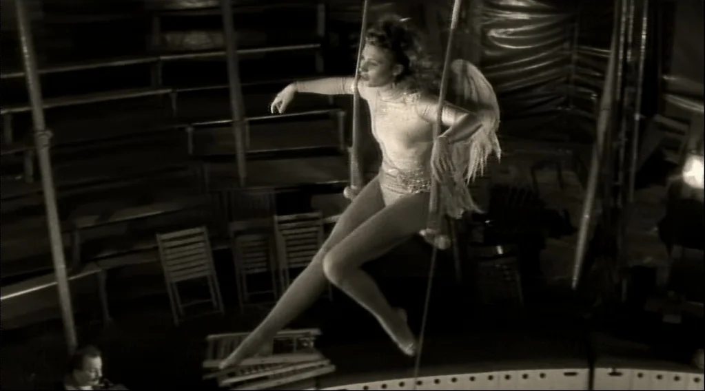 Wings Of Desire (1987) movie still: fullbody — A dark circus interior features wooden bleachers and metal support poles in the background. A woman…