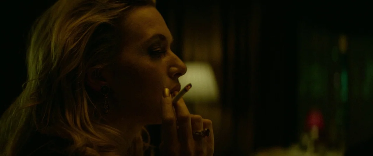 Triple 9 (2016) movie still: closeup — A dimly lit interior space with a soft glowing lamp in the background. A woman in profile smokes a cigarette…