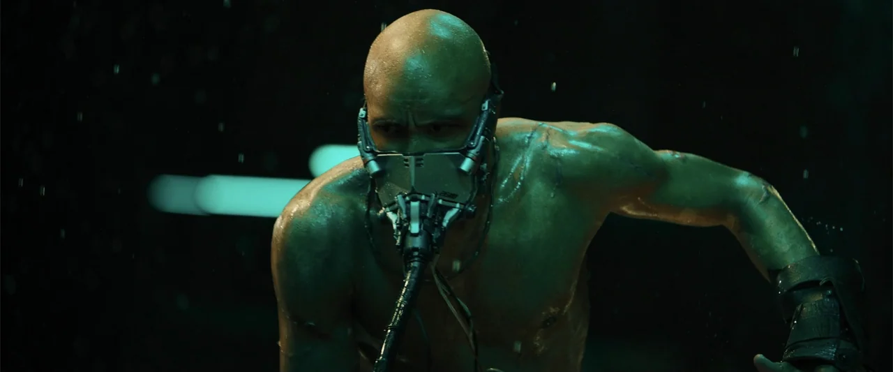 Black Adam (2022) movie still: medium — A dimly lit industrial interior features a dark, damp atmosphere with falling water droplets. A muscular,…