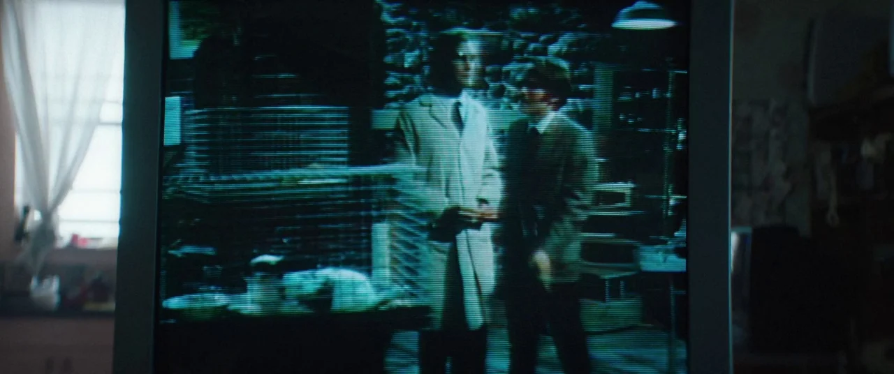 Broadcast Signal Intrusion (2021) movie still: medium — A low-resolution television screen displays a scene of two men in a dark room. One man in a…