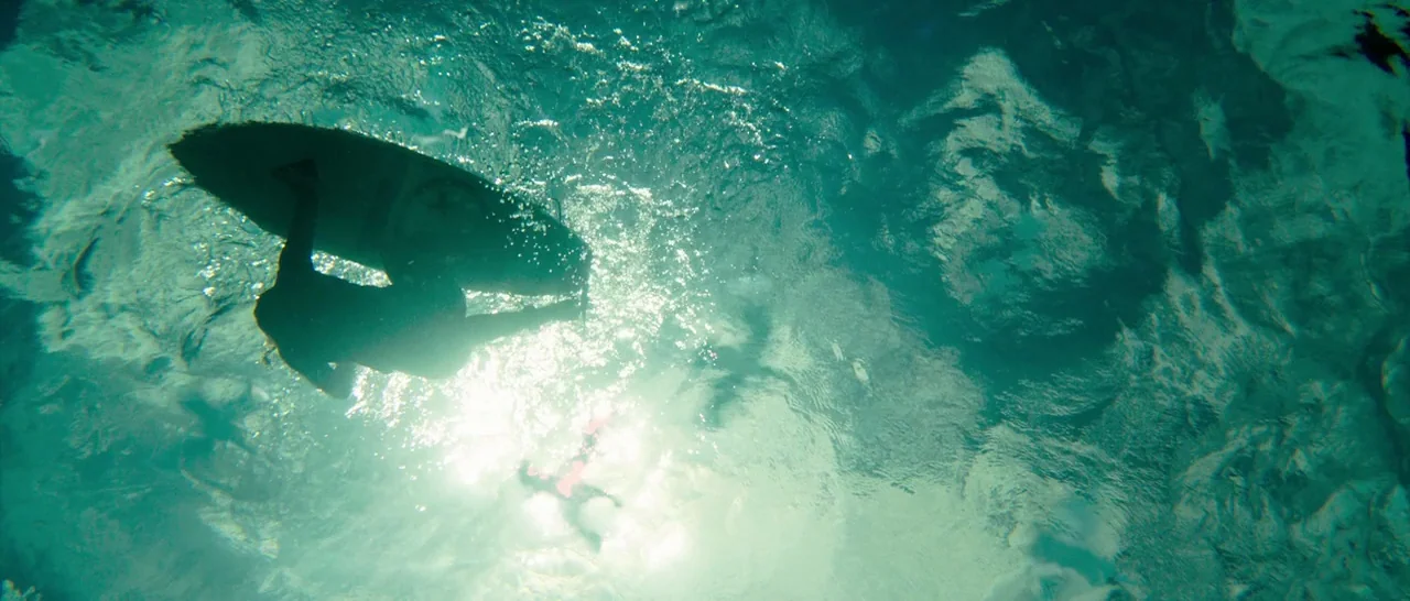 The Shallows (2016) movie still: wide — Sunlight penetrates the surface of clear blue water in the ocean. A lone surfer silhouette floats beneath a…