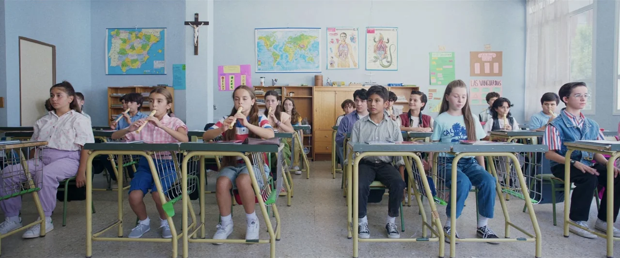 Voy a pasármelo bien (2022) movie still: medium — A 1990s school room features wooden desks arranged in rows under wall maps and posters. Multiple…