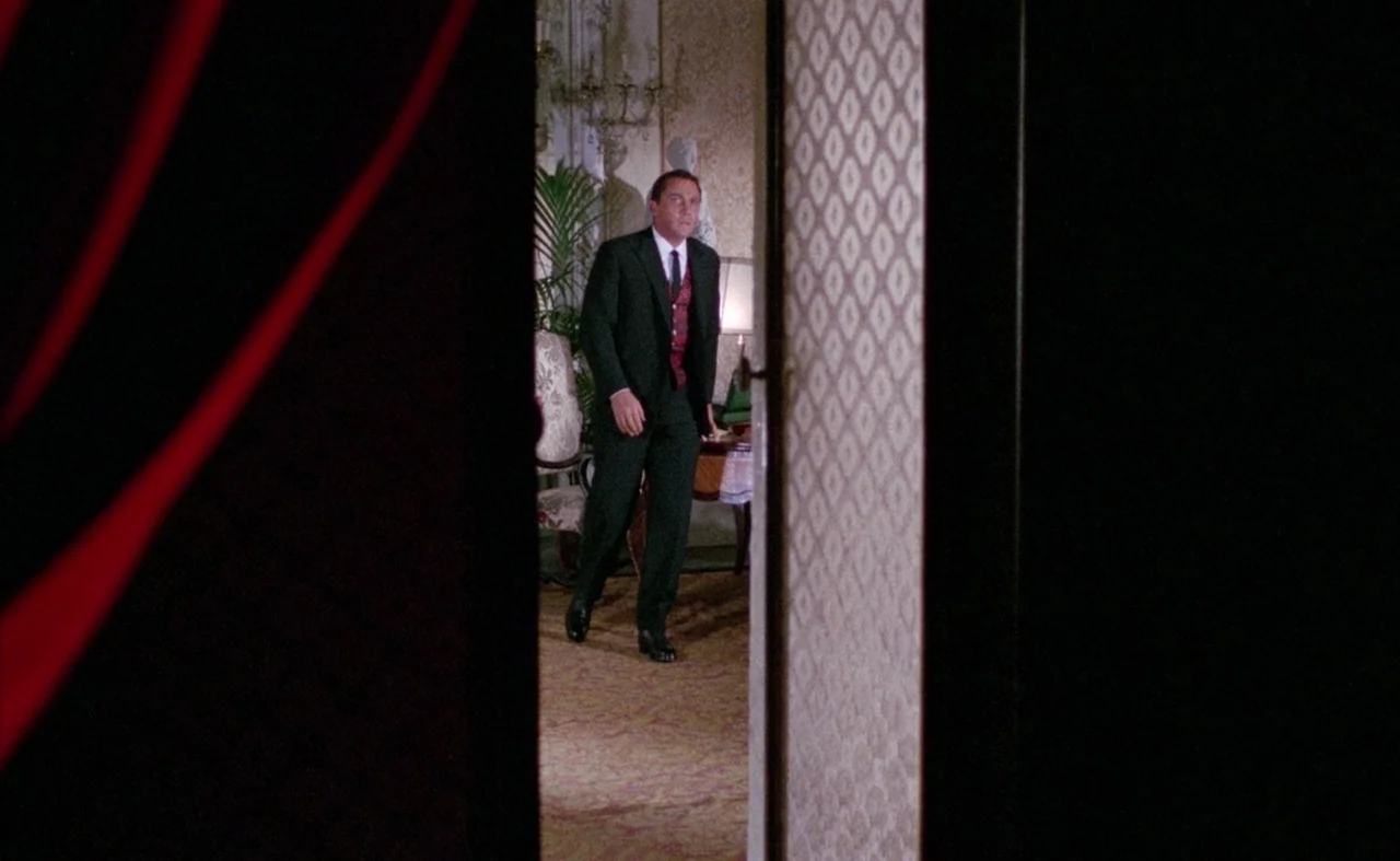 Blood And Black Lace (1964) movie still: medium — An opulent, vintage parlor features ornate wallpaper and a large indoor palm plant. A man in a…